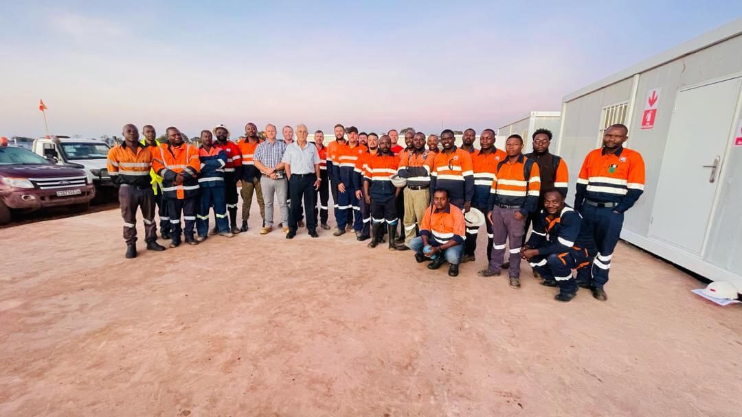 A large group of construction workers wearing orange and blue safety uniforms stand together in front of portable site buildings on a dirt surface. The sky is clear with a gradient from blue to light pink, indicating early morning or late afternoon.