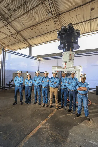 A group of eight people in blue uniforms and hard hats stands inside a large industrial warehouse. They are standing in front of considerable industrial equipment, with significant lighting coming from behind. The warehouse has a high, arched ceiling.