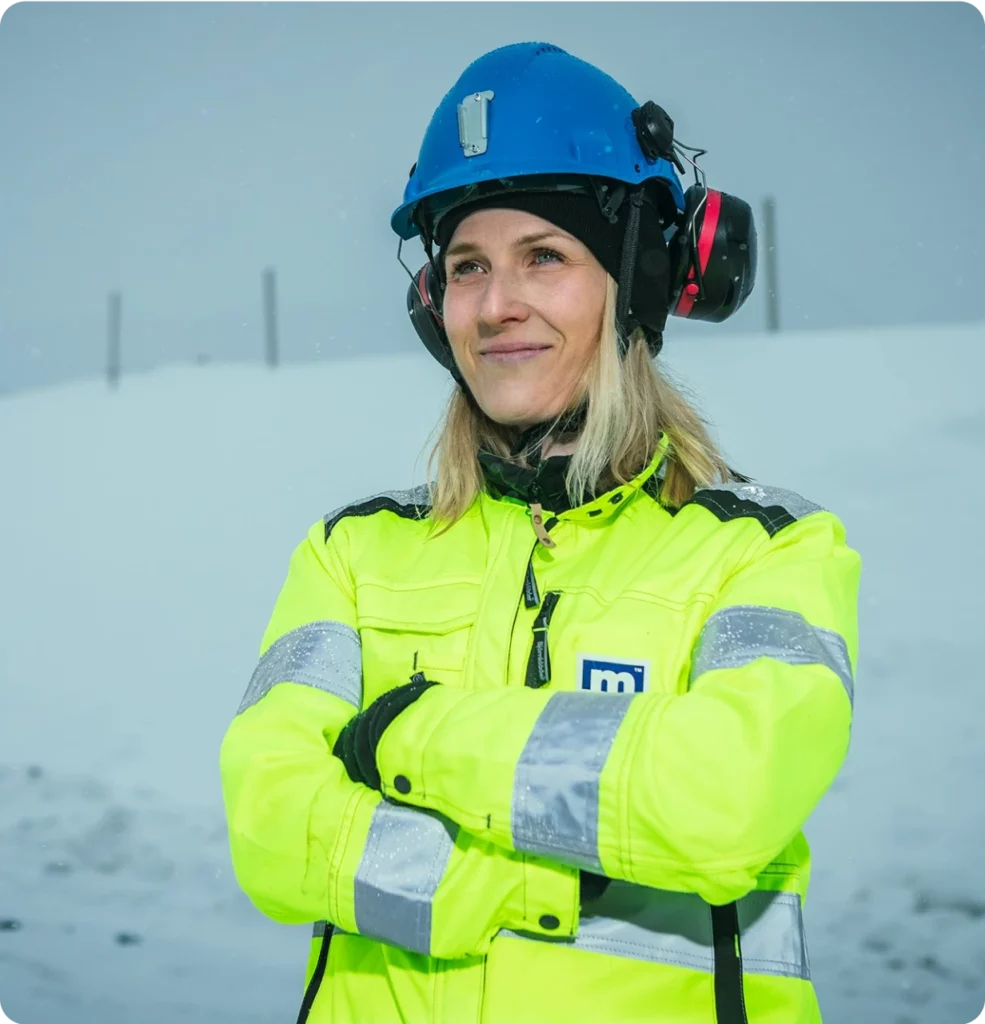 A person wearing a blue hard hat, ear protection, and a bright yellow high-visibility jacket stands with arms crossed in a snowy, outdoor environment. The individual is smiling and appears to be at a work site.