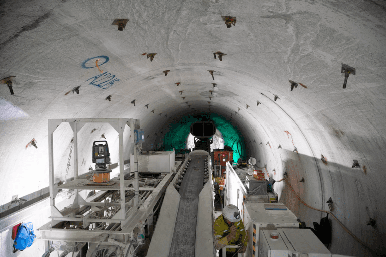 A wide tunnel under construction features various heavy machinery and equipment. Workers in safety gear are seen within the tunnel, focusing on their tasks. The tunnel wall is marked with some graffiti and is well-lit, giving a clear view of the construction process.