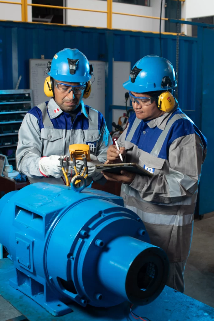 Two industrial workers in blue helmets and safety gear inspect and record information on a large electric motor inside an industrial facility. One is using a measuring device while the other writes on a clipboard. Both wear ear protection and safety glasses.