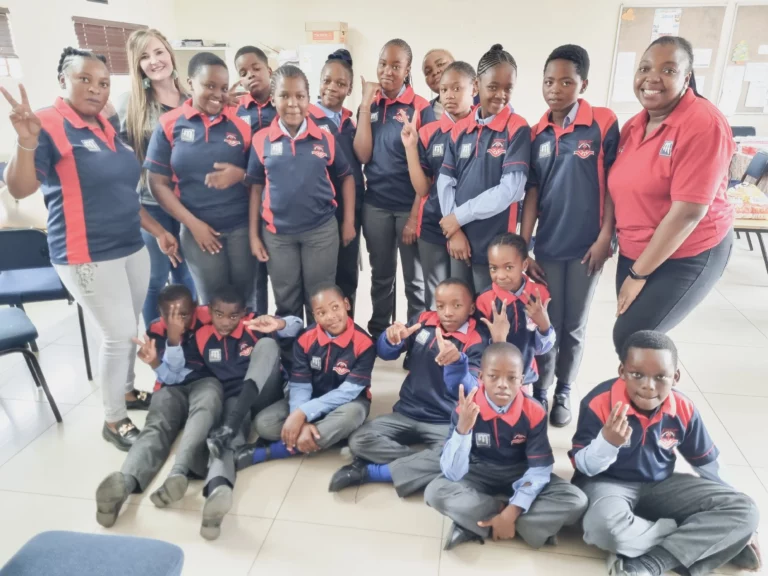 A group of students and two adults pose for a photo in a classroom. The students are wearing matching navy blue and red uniforms, with many making peace signs. The adults, one in jeans and a white shirt, the other in a red shirt, stand smiling with the group.