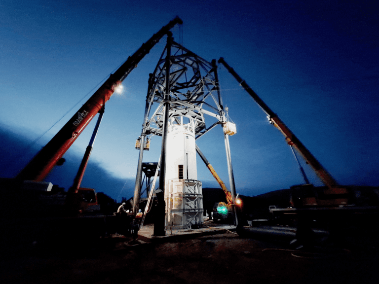 A group of cranes erects a large metal framework during dusk. The sky is dark blue, and the cranes' lights illuminate the structure and surrounding area. Workers and equipment are visible at the base of the framework.