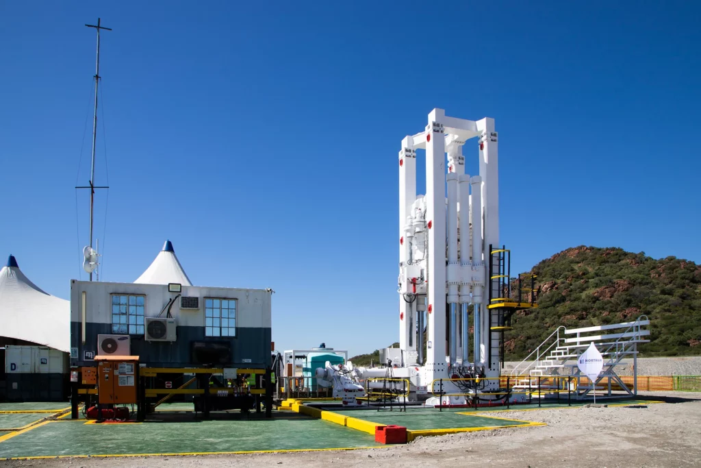 A white rocket stands vertically on a launch pad outside a launch facility, with mountains in the background. Nearby are tents, a wheeled building, and various equipment under a clear, blue sky. Steps lead to the base of the rocket, indicating preparation for launch.