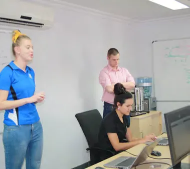 Three people are in an office setting. A woman in a blue shirt is standing and speaking, a man in a pink shirt is standing with arms crossed, and a woman in a black shirt is seated at a desk using a laptop. A whiteboard is visible in the background.