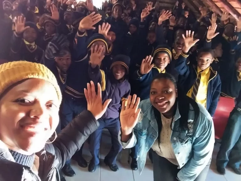 A diverse group of children in school uniforms enthusiastically wave at the camera. Two women in the foreground, also smiling and waving, appear to be taking a selfie with the children. The setting looks like a classroom or assembly hall.