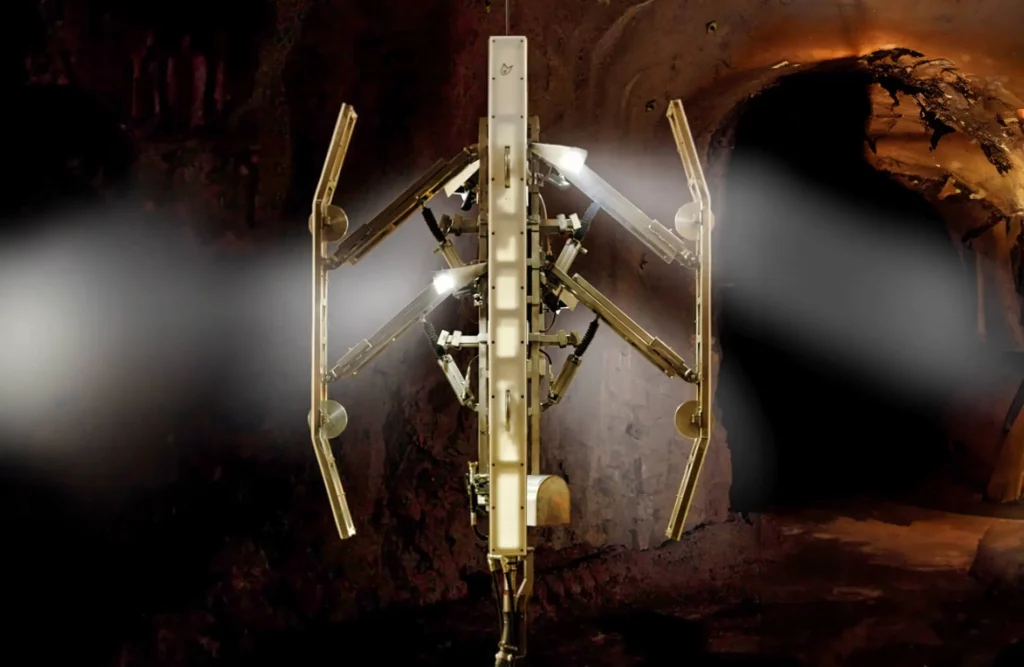 A vertical, mechanical device with multiple arms and jointed segments is illuminated against a dark, cave-like background. The machine appears to be a complex tool or robot, with lights on its arms shining onto the surrounding rocky environment.