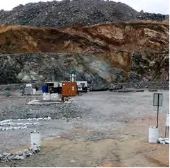 A rocky quarry with a backdrop of a large rock wall. The ground is covered in gravel and there are several construction materials and equipment scattered around, including barrels, a truck, and a portable office unit. The area appears to be an active work site.