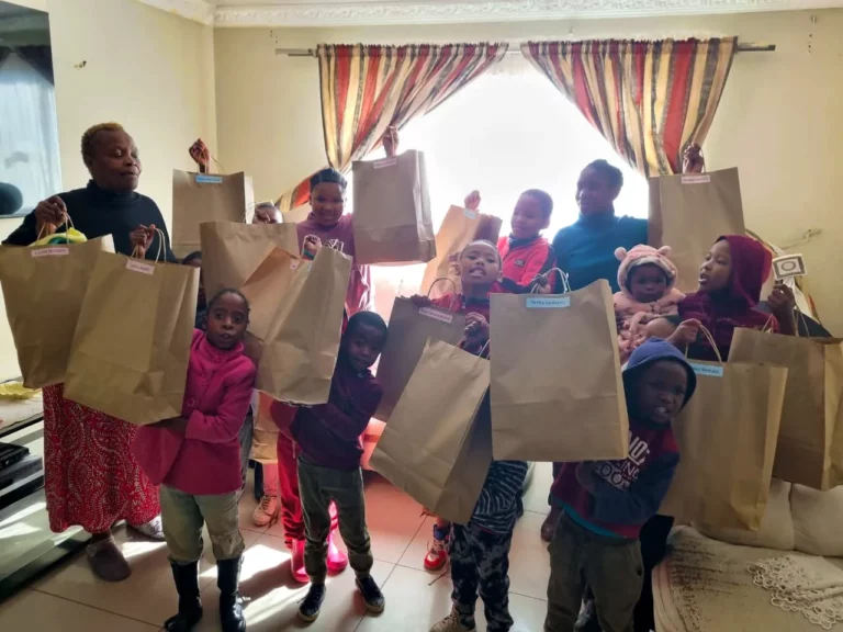 A group of smiling children and an adult stand in a well-lit room, holding brown paper bags with blue labels. The children are raising the bags enthusiastically. Colorful curtains hang by a window in the background, letting in natural light.