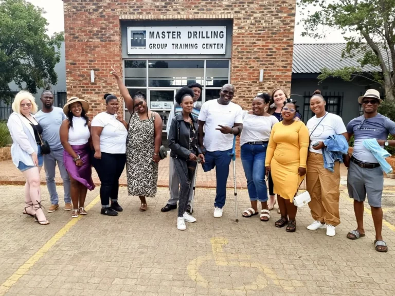 A diverse group of 14 people posing cheerfully in front of a brick building labeled "Master Drilling Group Training Center." They are standing on a pavement with a yellow-stripped handicapped parking space. Most are smiling and a few are striking playful poses.