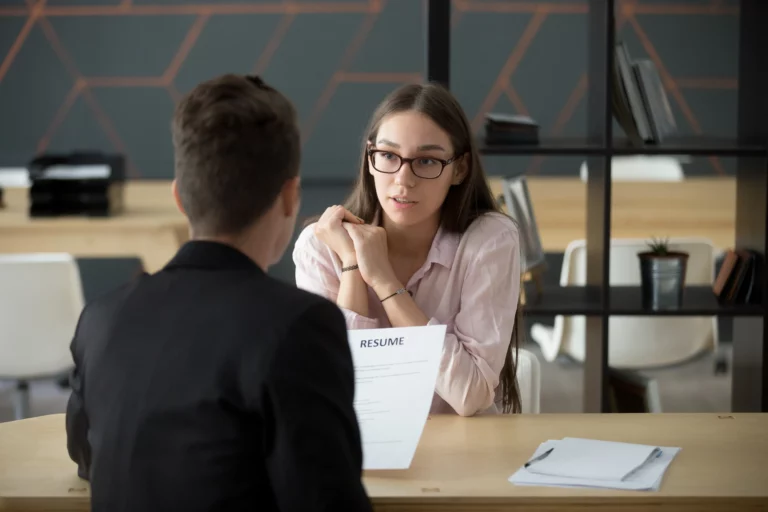 A woman with long hair and glasses sits across from a man in a suit at a table, holding her chin in her hands. The man faces away from the camera and holds a resume. The background shows a modern office setting with shelves and a geometric wall design.