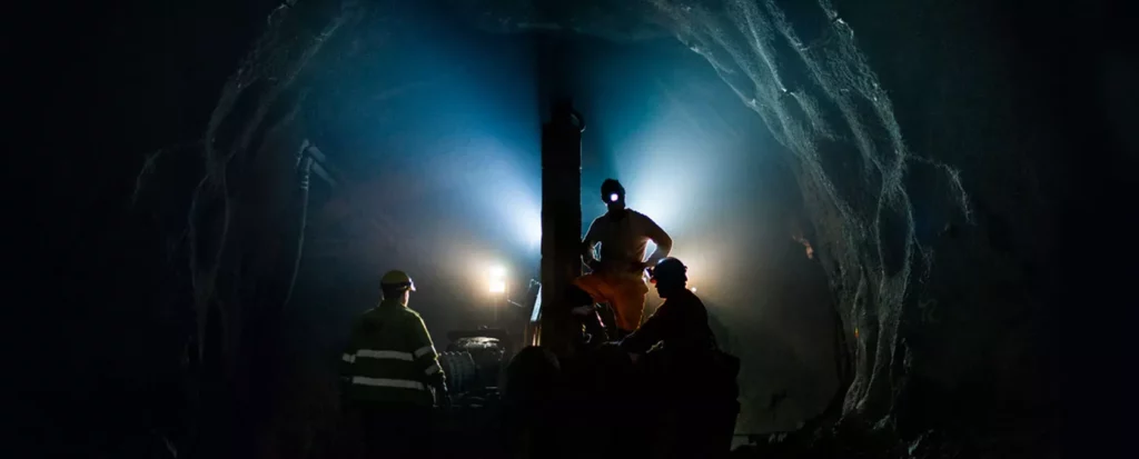 Three miners in safety gear work underground in a dimly lit tunnel. One miner stands and operates machinery illuminated by blue lights, while two others observe. The rocky tunnel walls encompass the scene, adding to the sense of depth and enclosed space.