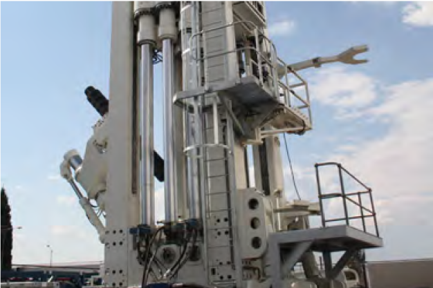 A large industrial drilling machine with multiple vertical metal cylinders and platforms is set against a background of a partly cloudy sky. The machine is accompanied by ladders and various attachments, showcasing its complex design and heavy-duty construction.