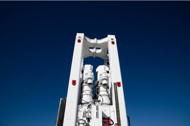 A close-up view of a white industrial machine component set against a clear blue sky. The component features two cylindrical elements in the center, framed by a rectangular structure with multiple red indicators.