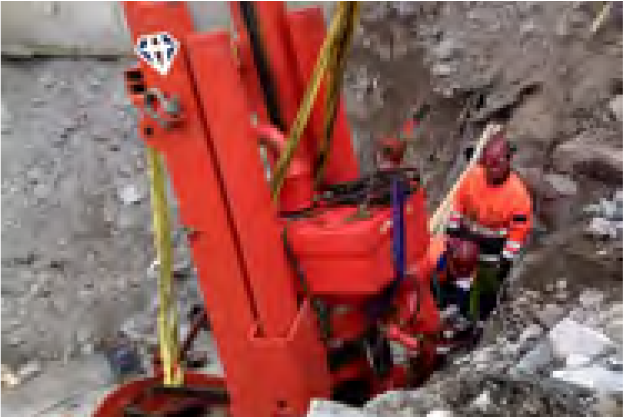 A worker in a safety vest operates a large, red construction drill amidst a rocky excavation site. Surrounding the worker and machine is a grey, rough terrain, suggesting ongoing construction or geological work.