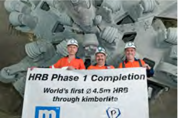 Three workers in orange safety uniforms and white hard hats are holding a banner that reads "HRB Phase 1 Completion. World's first Ø 4.5m HRB through kimberlite." They are standing in front of a large piece of industrial machinery with gears and components.