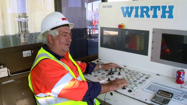 A worker in a white helmet and orange safety vest operates a control panel labeled "WIRTH." He sits inside a control room with monitors and various buttons on the panel. A can of soda is placed on the panel's edge.