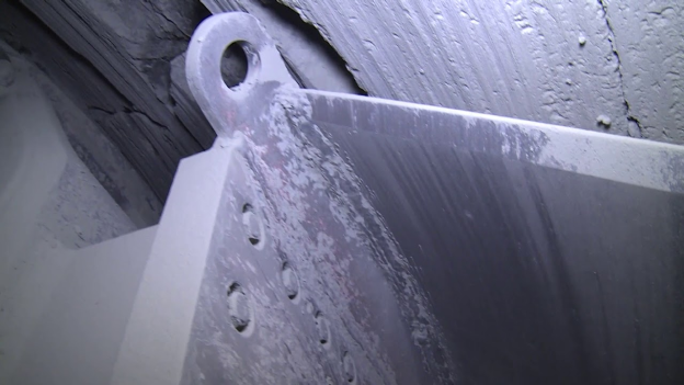 Close-up of a worn and dusty metallic surface with a holed attachment point, possibly part of industrial equipment or heavy machinery. The background features a rough, dimly-lit surface with streaks of dust or residue, indicating a likely underground or cave environment.