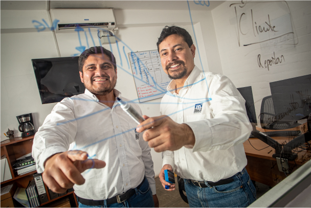 Two men in white shirts are smiling and collaborating at an office. One of them is writing on a transparent glass board, pointing out details highlighted in blue marker. Office amenities such as a TV, coffee maker, and a bookshelf are visible in the background.