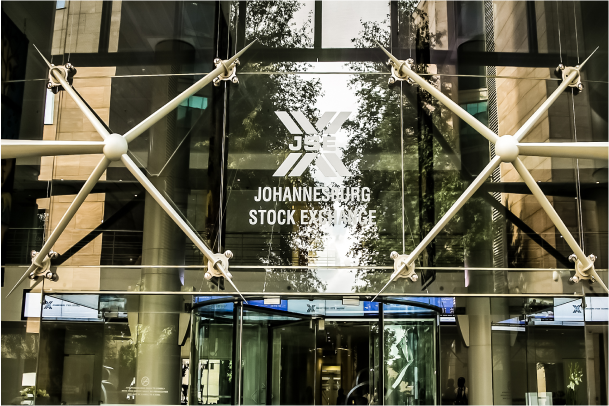 The entrance to the Johannesburg Stock Exchange (JSE) building, featuring glass doors and large metal supports. The JSE logo and name are prominently displayed on the glass facade. Trees and reflections are visible through the glass.