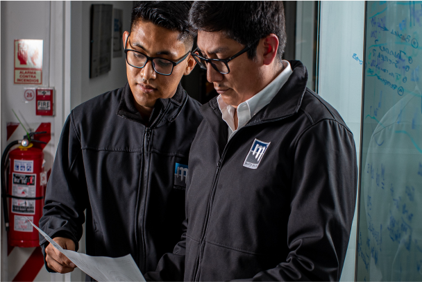 Two men in matching dark jackets, both wearing glasses, closely examine a piece of paper. They stand next to a wall with writing on it, possibly in a workplace setting. A fire extinguisher and emergency sign are visible in the background.