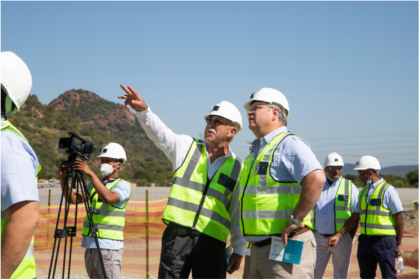 A group of construction workers wearing hard hats and reflective vests stand at a construction site. One man, pointing into the distance, is speaking to another man. A cameraman records the scene, while others in the background appear to be observing.