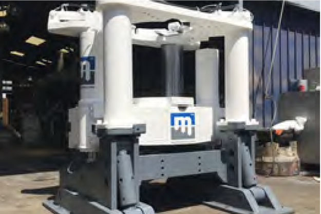 A large hydraulic press machine with white upper and gray lower parts is positioned indoors near a blue vertical storage tank. The press features the distinctive logo of its manufacturer on its front panel. The background shows a workshop area.