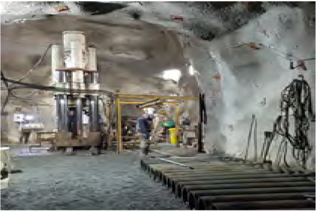 Miners work inside an underground mine with heavy machinery and equipment. The environment is rocky, with a large drill and conveyor system visible. Cables run along the walls and ceiling, and a few workers are engaged in various tasks.