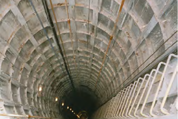 A deep, cylindrical tunnel with a concrete, segmented interior and descending metal ladder on the right side. Electric lights are spaced evenly along the tunnel, illuminating the interior walls and creating a vanishing point effect as the tunnel stretches into darkness.