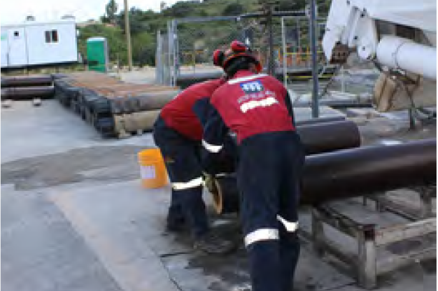 Two workers in red shirts, black pants, and safety helmets and gear are handling a large cylindrical pipe on a worksite. The background shows a porta-potty, a white trailer, and other equipment and materials. The setting appears to be an industrial or construction area.