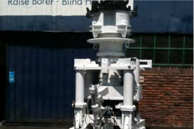 White industrial drilling machine with multiple pistons and mechanical components positioned in front of a blue door and a brick wall. Signage in the background reads "Raise Borer - Blind H.