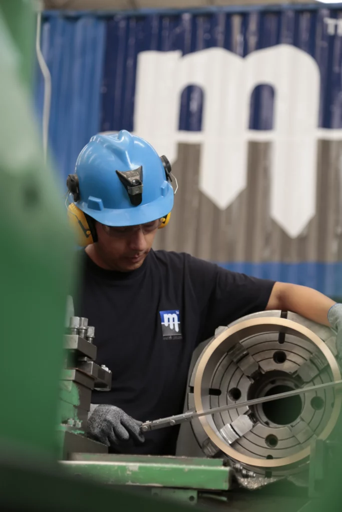 A worker wearing a blue protective helmet and yellow ear protection operates machinery in an industrial setting, focusing on a large circular metal component. He is wearing a black shirt with a small white and blue logo on the chest. A blurred blue and white backdrop is visible.