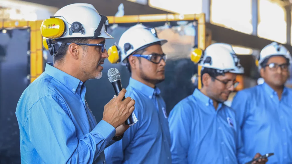 Four men in blue shirts and white safety helmets are standing in a line. The man on the far left is speaking into a microphone, while the others listen attentively. All are wearing safety glasses and appear to be in an industrial or construction setting.