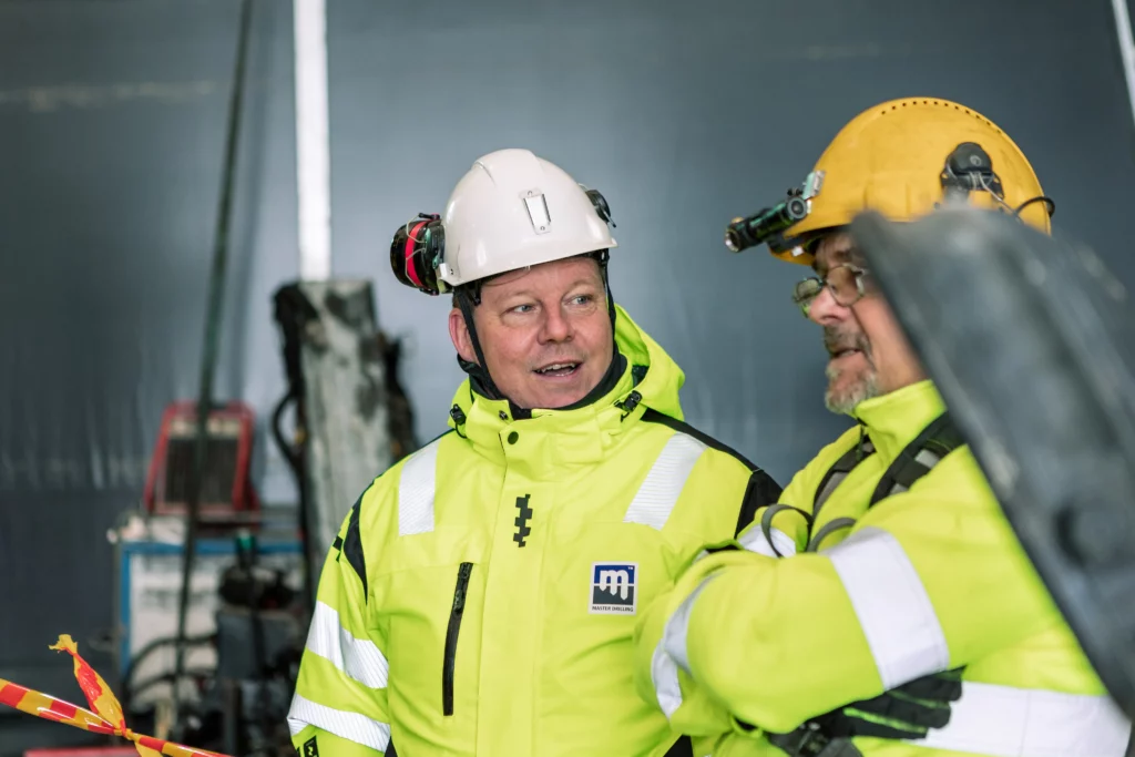 Two construction workers wearing high-visibility jackets and hard hats stand indoors, engaged in conversation. One has a yellow hard hat with a headlamp, and the other has a white hard hat. They appear to be discussing a project in an industrial setting.