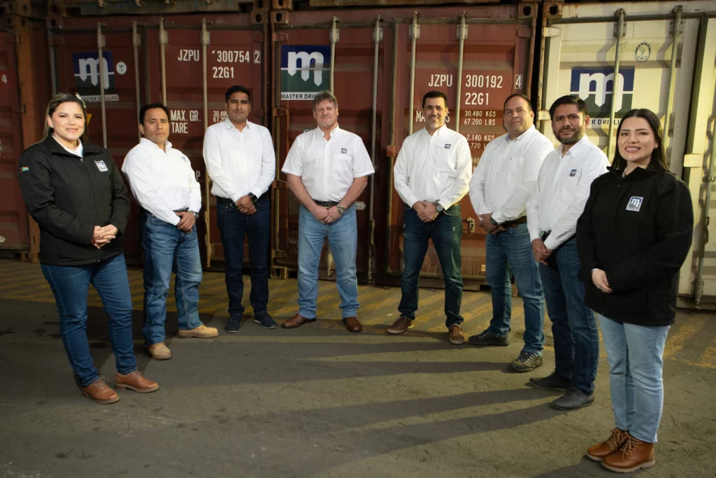 A group of eight people dressed in white shirts and jeans stand in front of shipping containers, smiling for a photo. Two individuals on the left and right sides wear black jackets. The containers in the background display various identification numbers and logos.