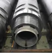 Close-up of a large, cylindrical industrial component with threaded ends and patterned grooves on the outer surface. The object appears to be a section of heavy-duty piping or machinery, with additional similar components in the background.