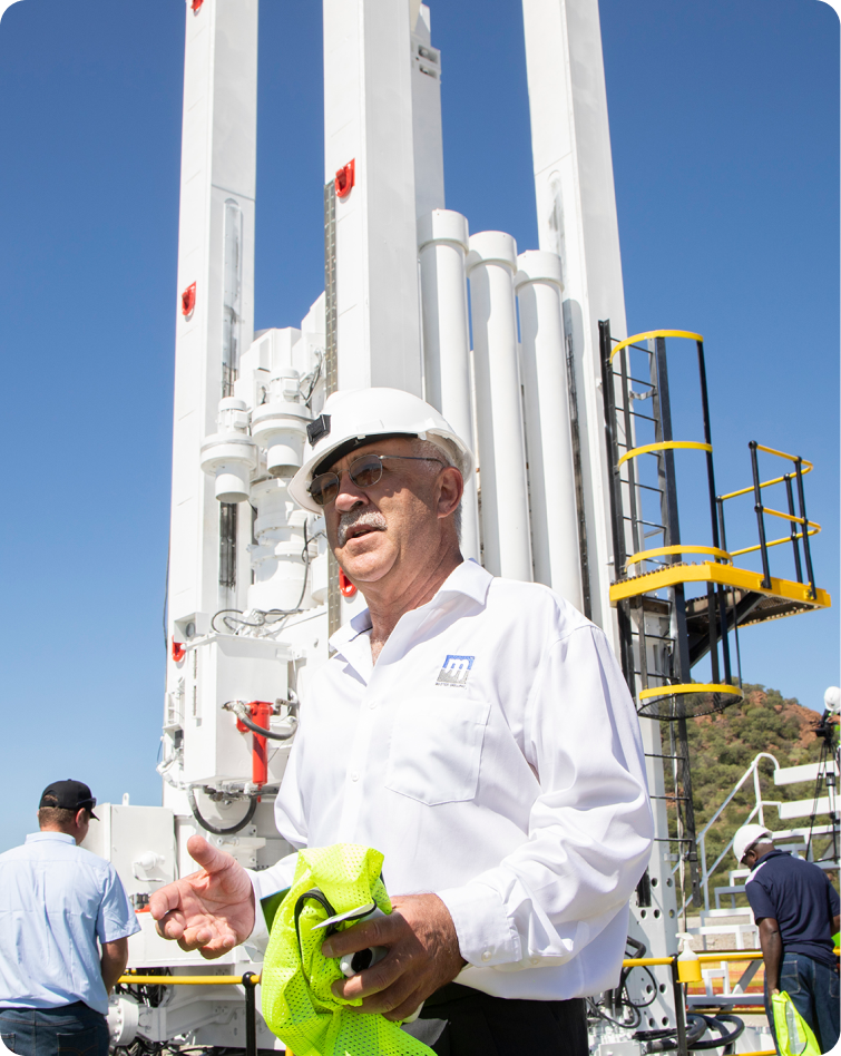 A worker in a white shirt with the logo "M" and a white hard hat stands in front of industrial equipment and a tall machine. He holds a yellow safety vest. Other workers and a ladder structure with railings are visible in the background. The sky is clear and blue.