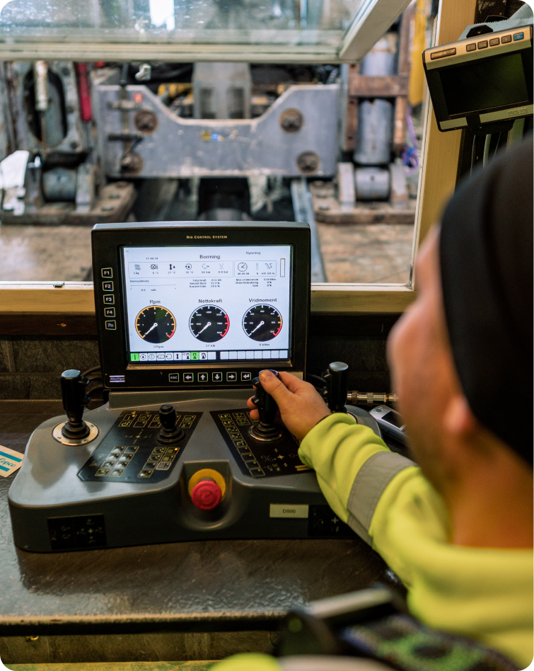 A person in a high-visibility jacket operates a control panel with multiple buttons, levers, and a screen displaying gauges and metrics. The back of machinery is visible through the window in front of the operator.