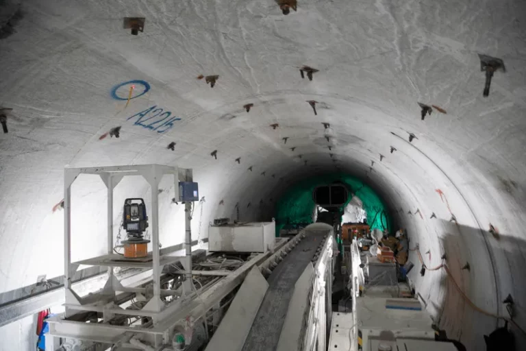 An interior view of a large tunnel under construction. The curved walls are lined with bolts and some writing. Construction equipment and machinery line the floor and sides. A green tarp partially obscures the tunnel’s end in the distance.