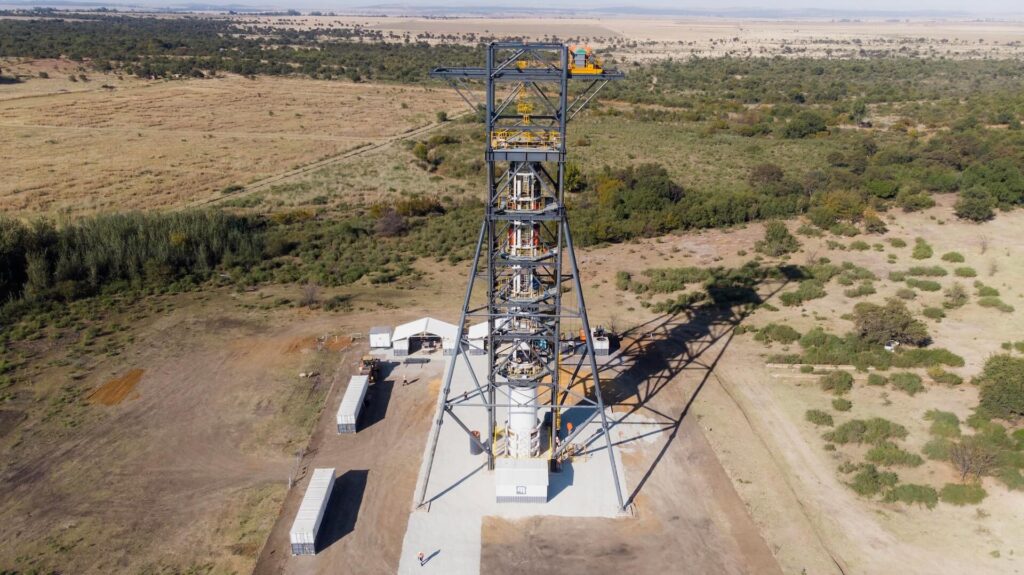 A tall steel lattice tower stands on a concrete foundation in a rural area. The structure is surrounded by sparse vegetation and an expansive landscape stretching to the horizon. Small structures and storage containers are positioned around the tower's base.