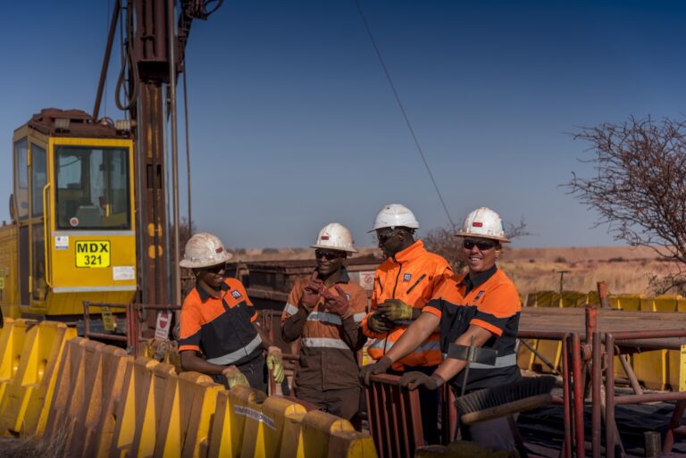 A group of four construction workers in safety gear share a light moment at a construction site. They are wearing orange and black uniforms, white helmets, and safety glasses. Behind them are yellow barriers, heavy machinery, and equipment under a clear blue sky.