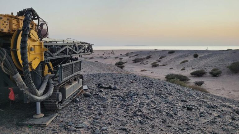 A large industrial machine with metal hoses and components stands on rocky terrain in a barren, desert-like landscape. In the distance, there is a flat horizon under a pastel-colored sky at dawn or dusk. Sparse bushes and rocks are scattered across the scene.