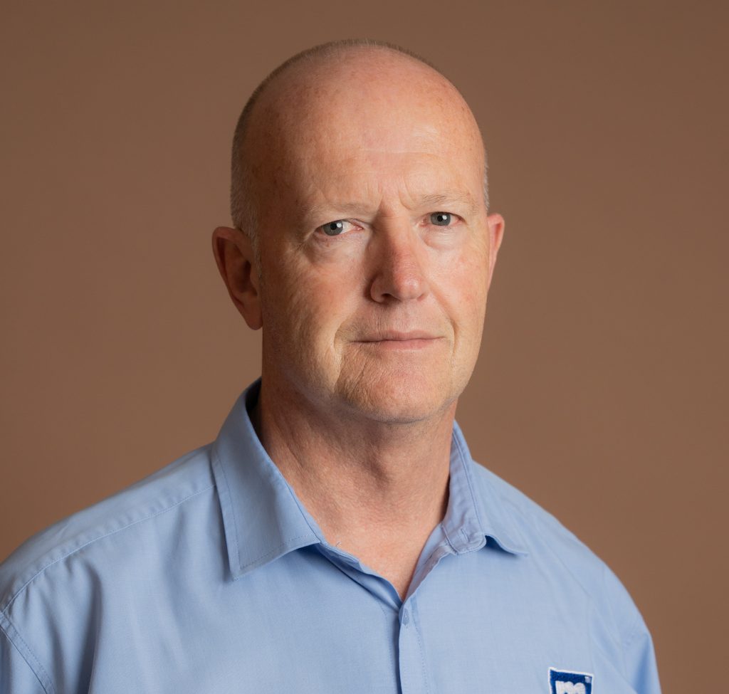 A middle-aged man with a bald head wearing a light blue collared shirt stands against a plain brown background, looking directly at the camera with a neutral expression.