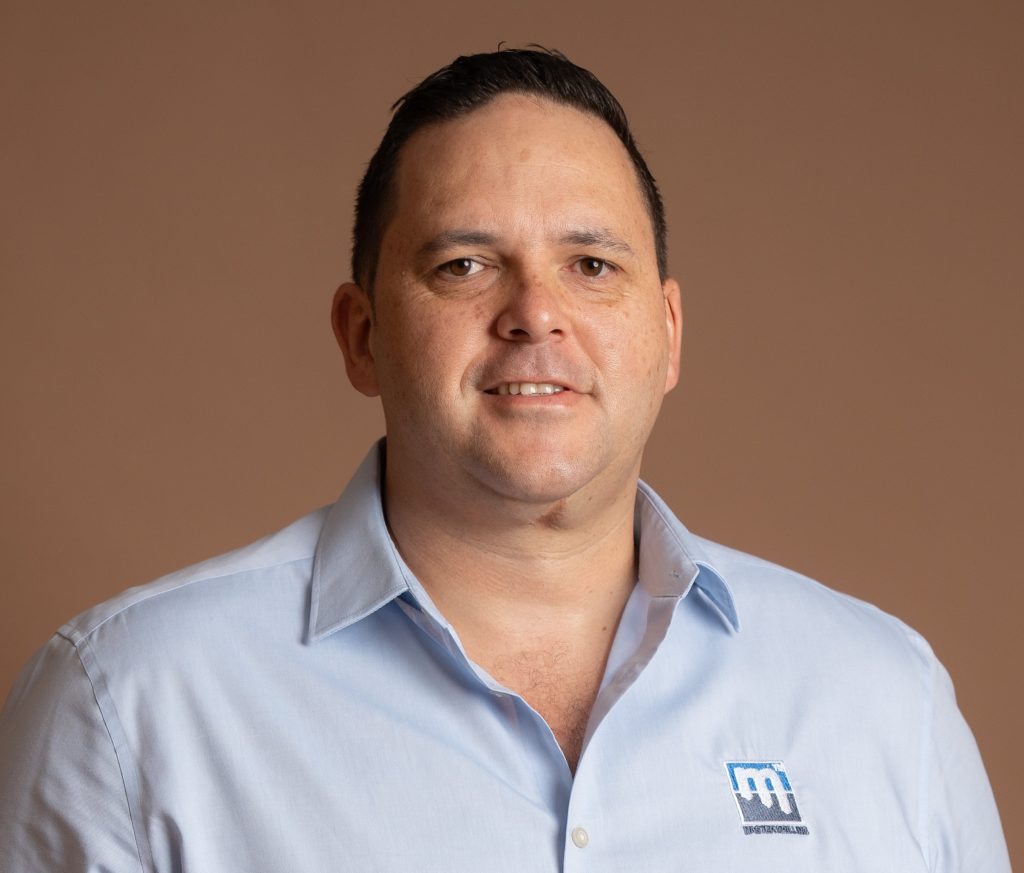 A man with short dark hair wearing a light blue collared shirt with a blue and white embroidered logo stands in front of a plain beige background, looking at the camera and smiling slightly.
