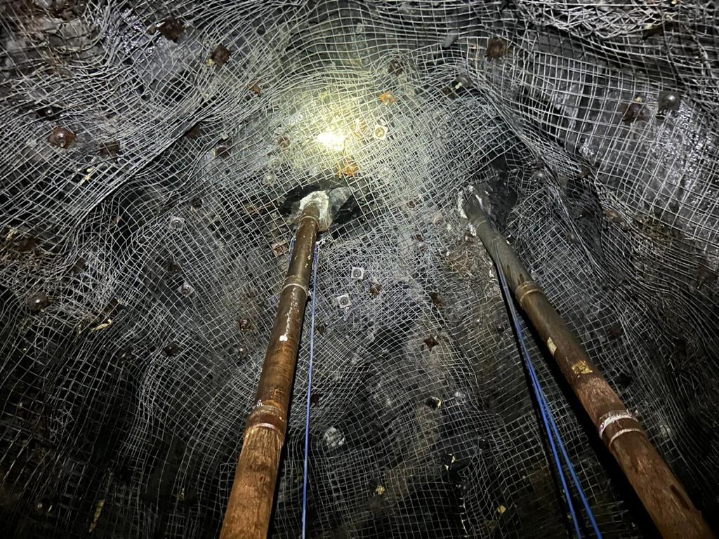 A rocky underground tunnel with a ceiling reinforced by wire mesh. Two wooden poles stand vertically, propping up the structure. A single bright light illuminates the scene, highlighting the dark, rugged surfaces.