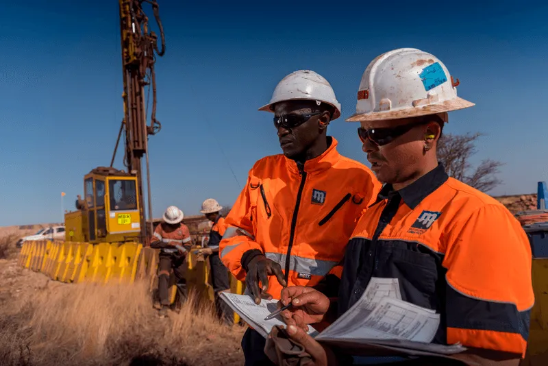 Two workers in orange and blue safety gear and white helmets discuss something while holding documents at a mining or construction site under a clear blue sky. A drilling rig is visible in the background, alongside other workers engaged in tasks.