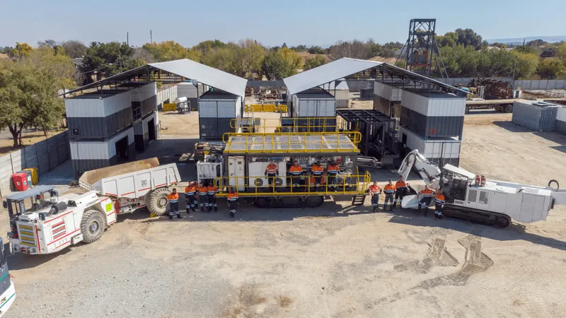 An overhead view of an industrial site featuring large machinery and equipment. Workers in orange safety gear are gathered in front of the equipment. The area is surrounded by several metallic buildings and trees can be seen in the background.