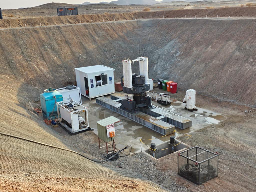 An industrial site set in a large excavation pit, featuring machinery, storage tanks, and a small control building. Various equipment and containers are organized on the concrete platform amidst a barren, desert-like landscape.