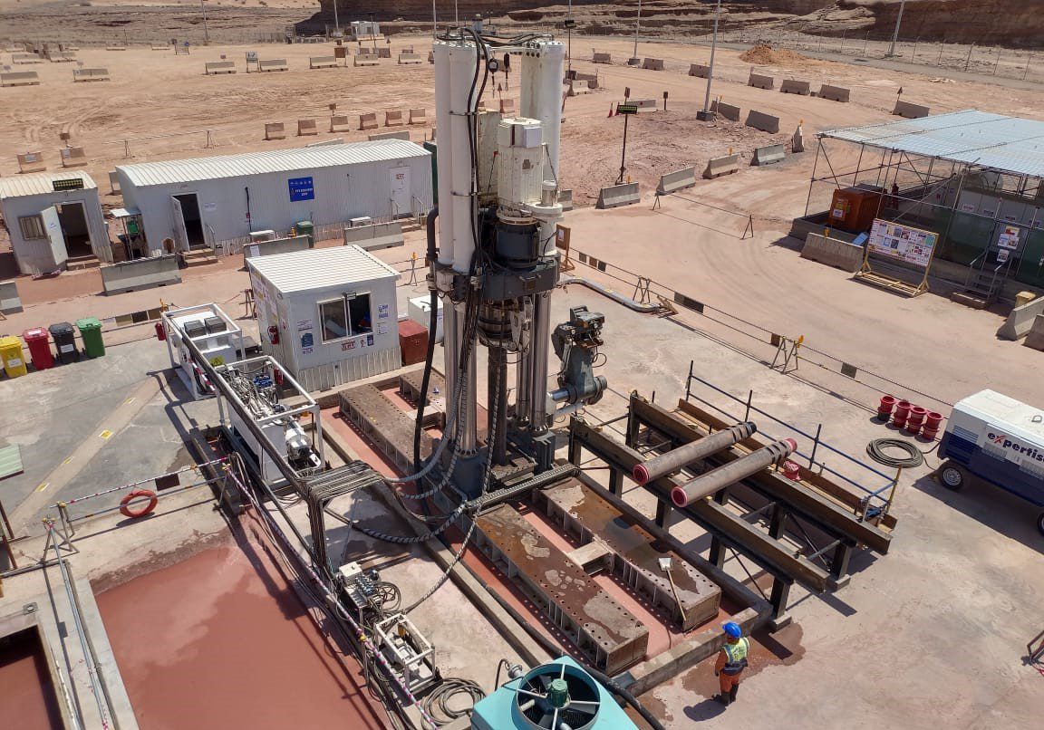 Aerial view of a construction site featuring a large drilling rig surrounded by metal pipes and industrial equipment. Containers and a temporary office building can be seen nearby. A person in a hard hat stands near the rig, with barren land in the background.