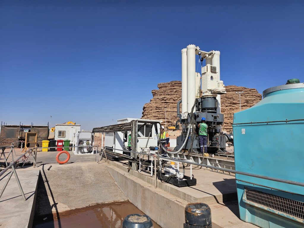 A construction site with machinery and workers in hard hats. The scene includes a large rock formation in the background, an industrial structure with pipes, and several containers. The sky is clear and blue.